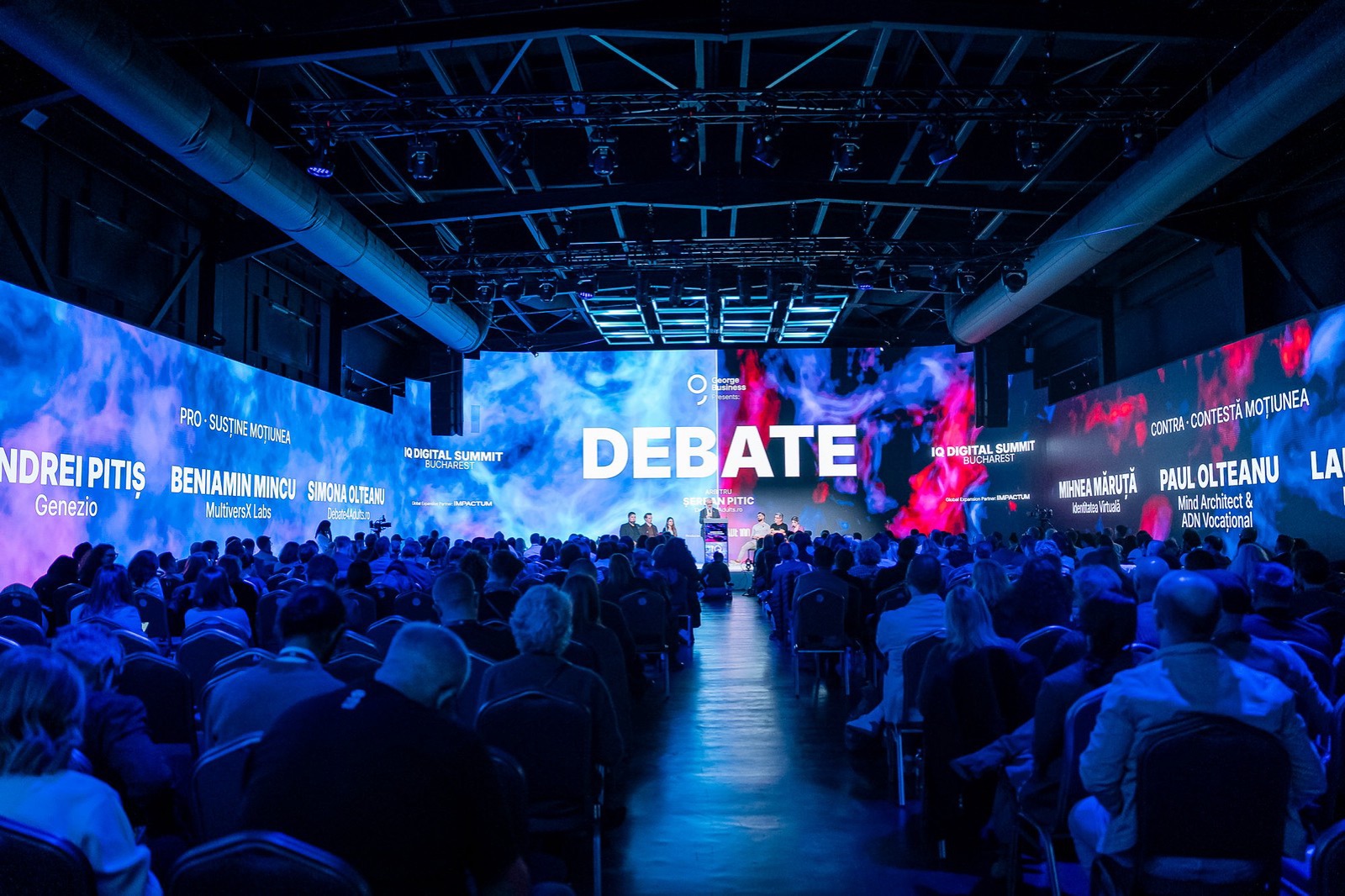 Sala plină la dezbaterea Oxford de la IQ Digital Summit, cu numele echipelor PRO și CONTRA pe ecrane